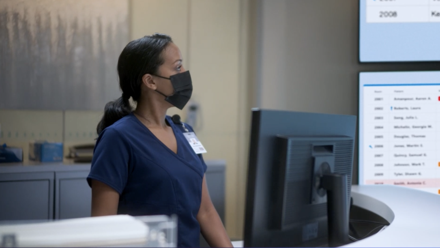 A masked healtcare worker stands at her station.