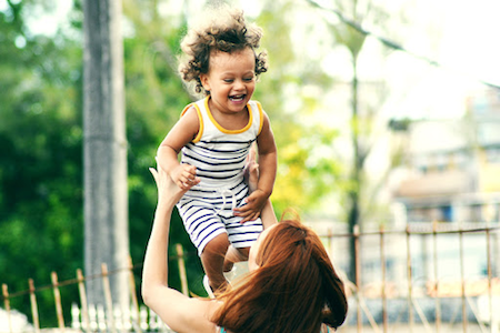 Caucasian female playfully tossing toddler in air. The two are smiling at each other and look unaware of camera.