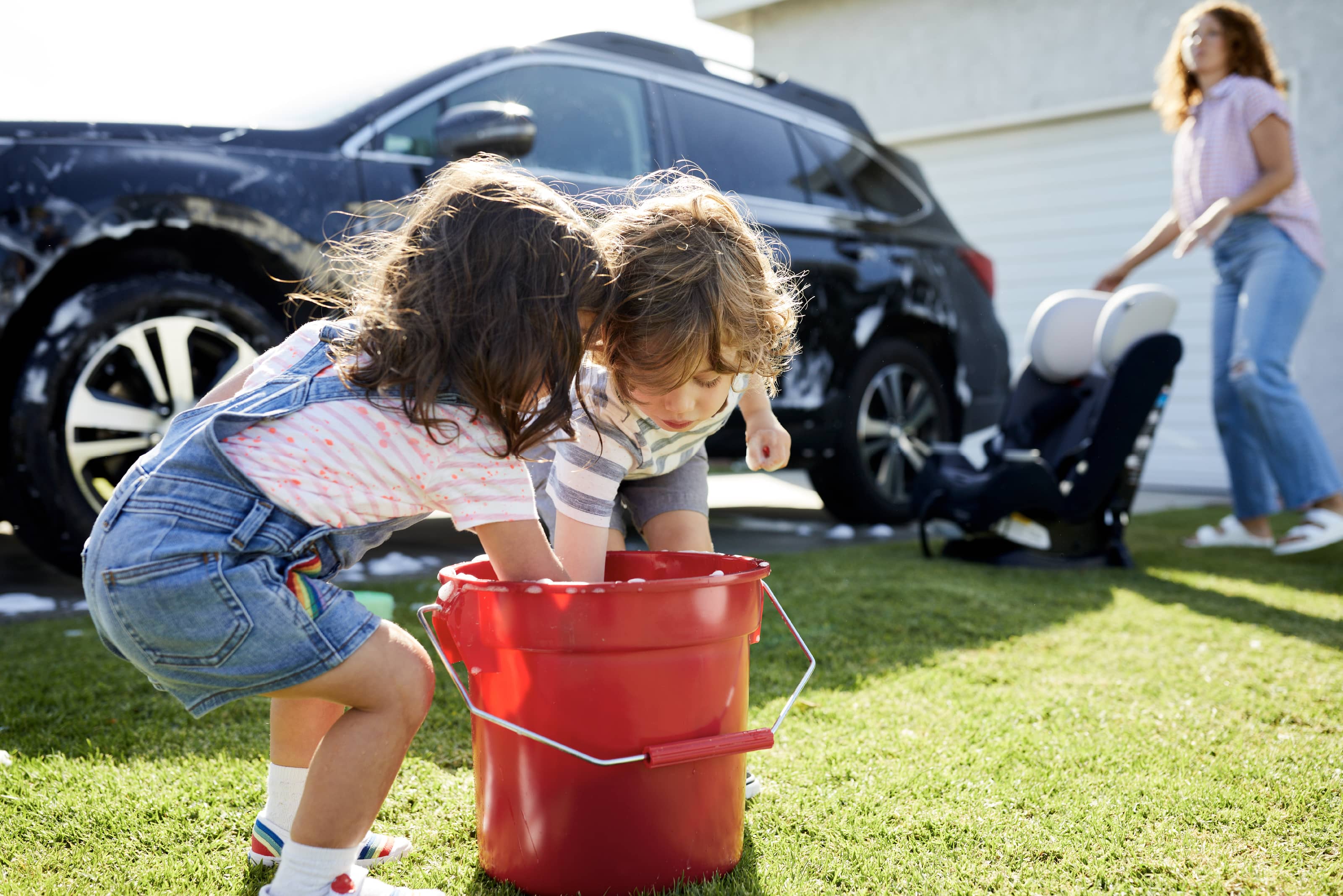 Two children reach into a bucket to continue washing a car. A woman moves a car seat.