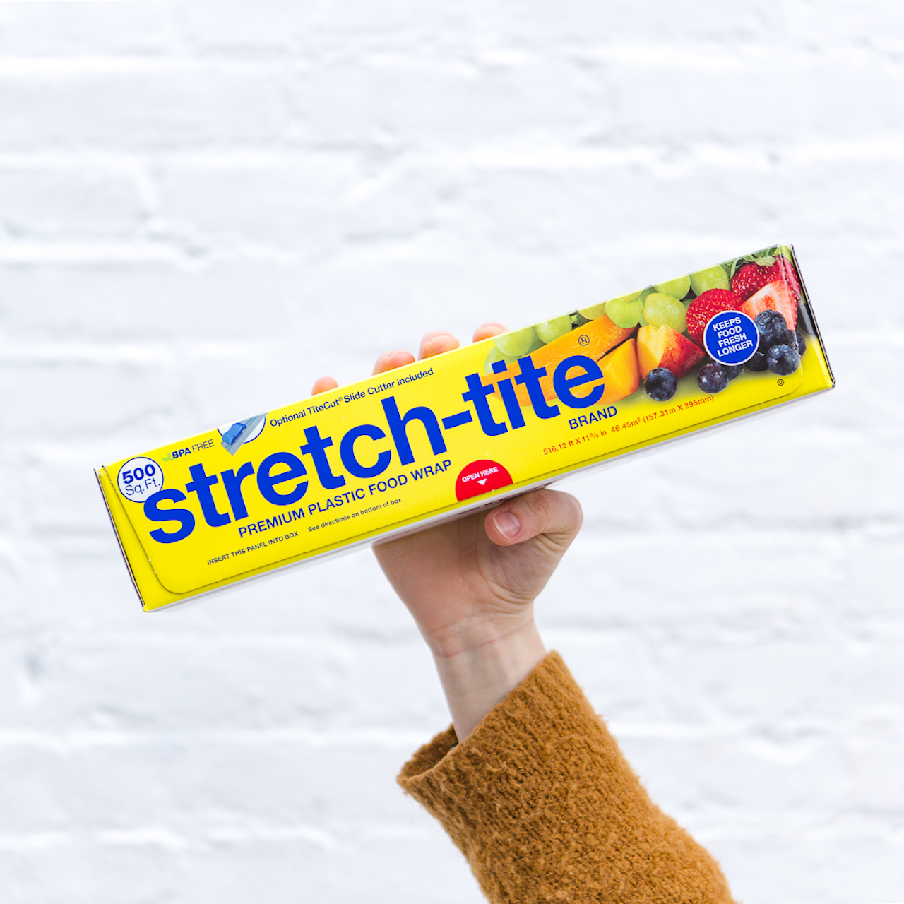 female hand holding a Rectangular box of Stretch-Tite plastic wrap in front of white brick wall