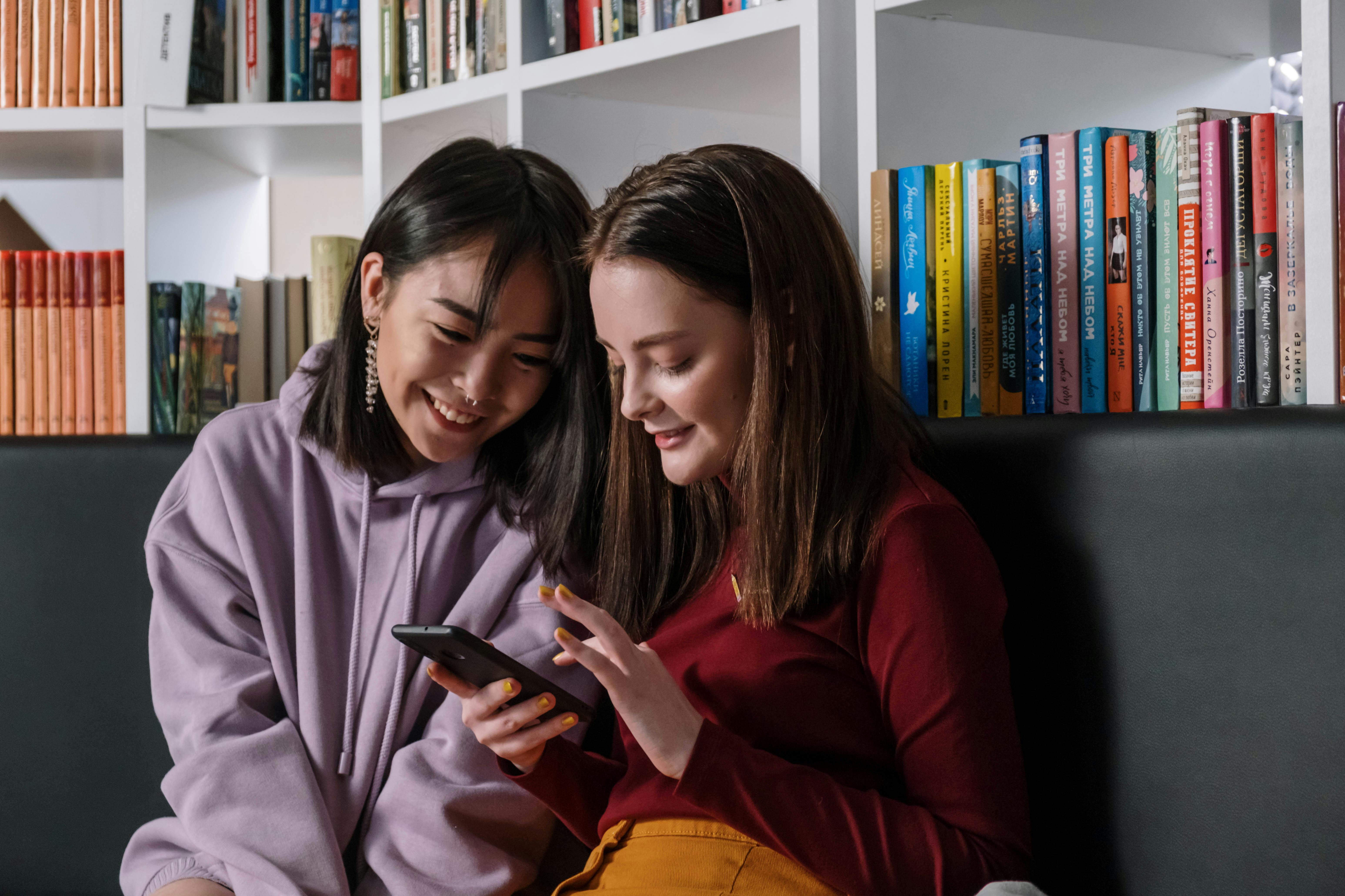 two teenage girls looking at phone on couch in front of a bookshelf