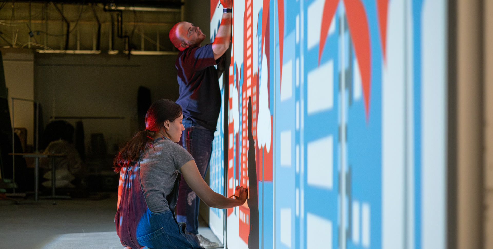 Two people paint a large mural on a wall, adding vibrant colors and geometric shapes.