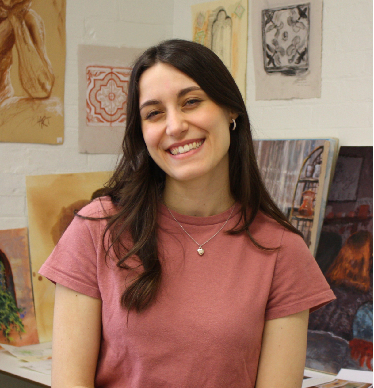  A young woman smiles, seated in front of artwork, wearing a casual pink shirt.