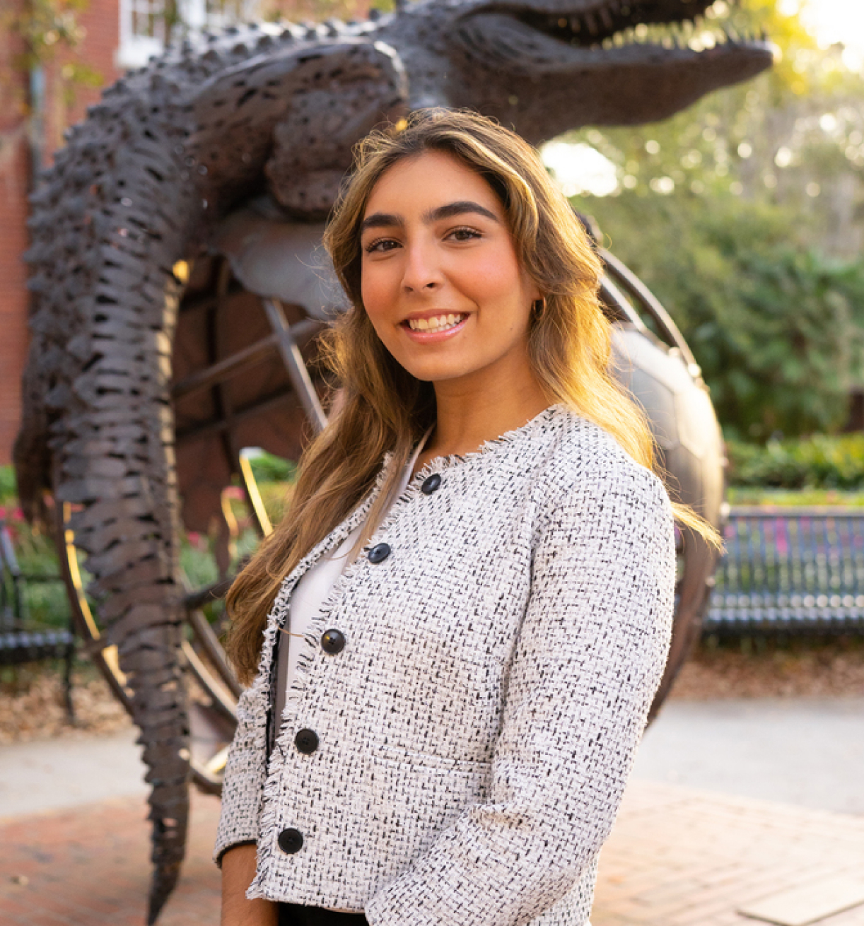 A smiling woman stands in front of an outdoor sculpture, wearing a textured jacket.