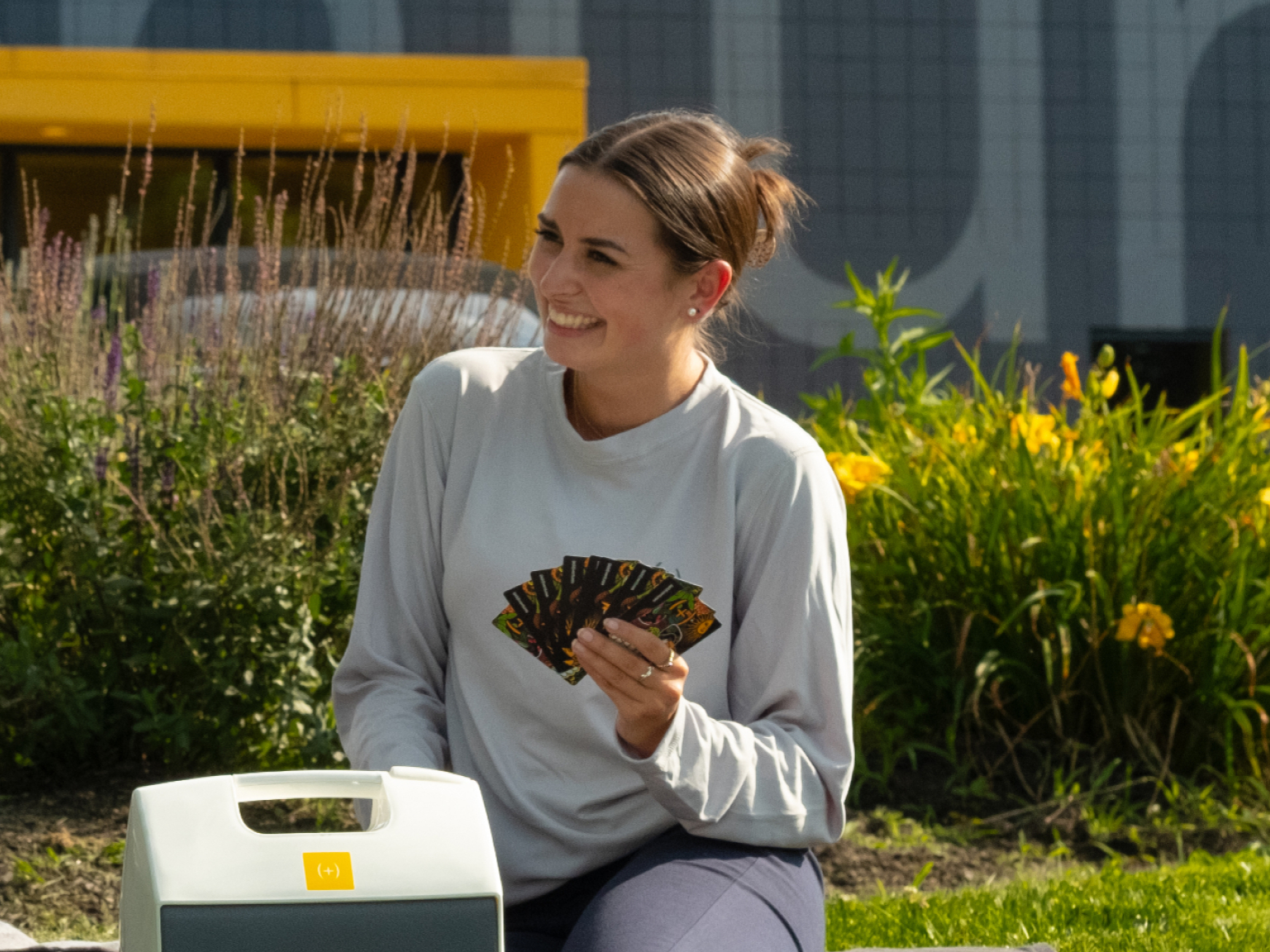 A person sits outdoors, holding a fan and smiling, with yellow flowers in the background.