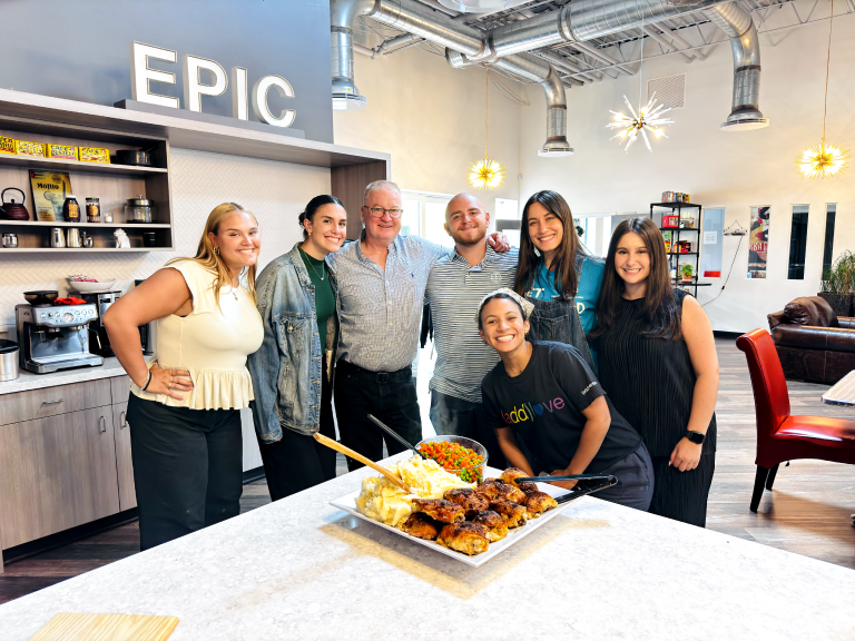 A group of six people stand together smiling in a kitchen area with a sign reading “EPIC” in the background, gathered around a table with food skewers displayed.