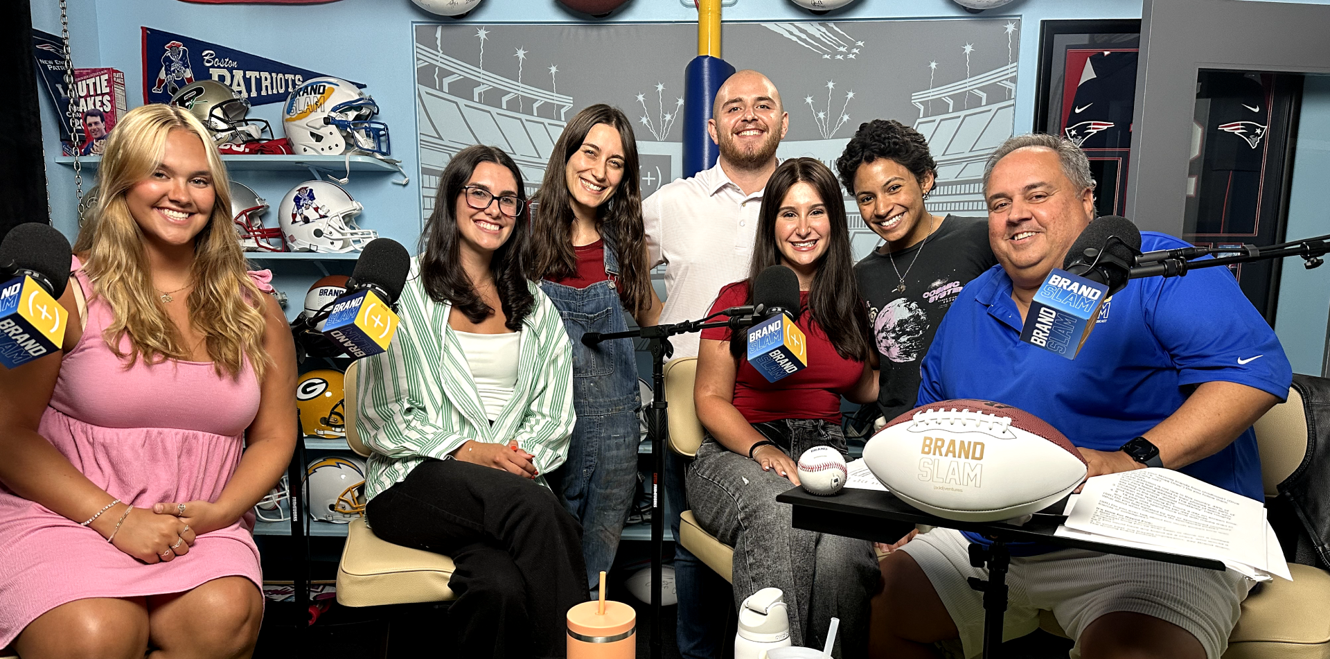 A group of seven people sit together in a studio setting, smiling at the camera. One person holds a football, and microphones are visible in front of several of them.