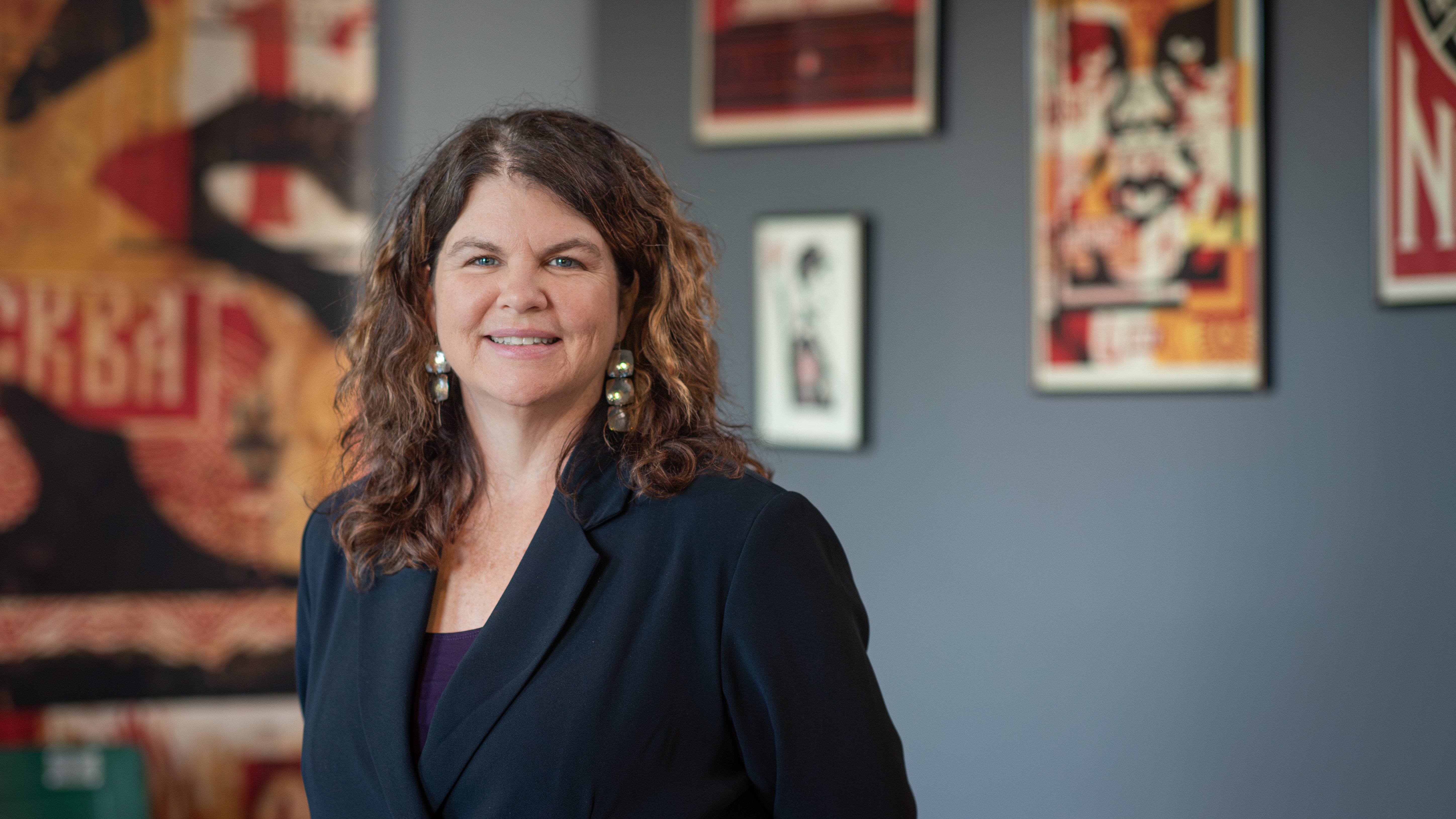 “Portrait of a woman in a blazer standing in front of framed posters on a gray wall.”