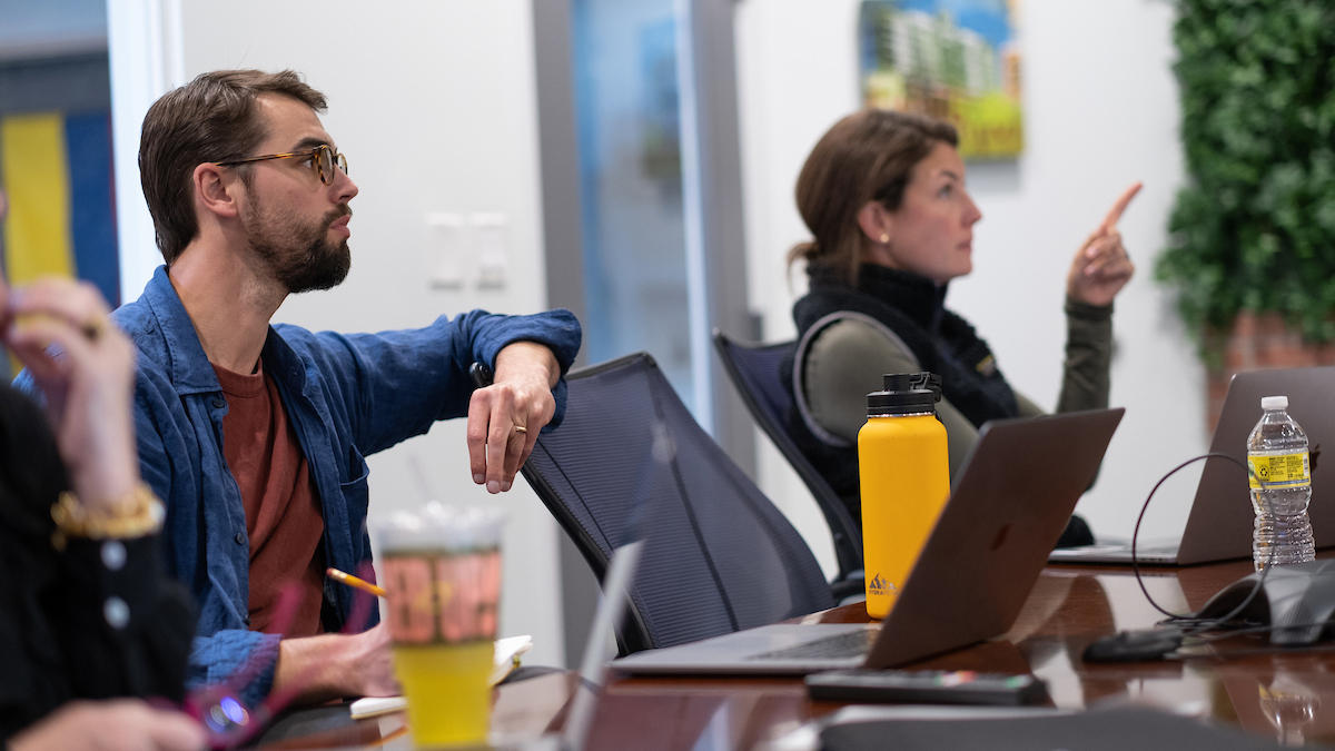 Two focused team members in a meeting, one gesturing while the other listens attentively, with laptops, notebooks, and water bottles on the table.