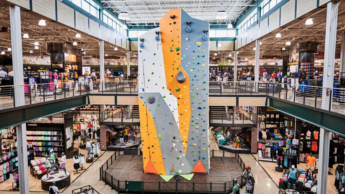 Retail space with a central rock climbing wall surrounded by shoppers and sportswear displays.