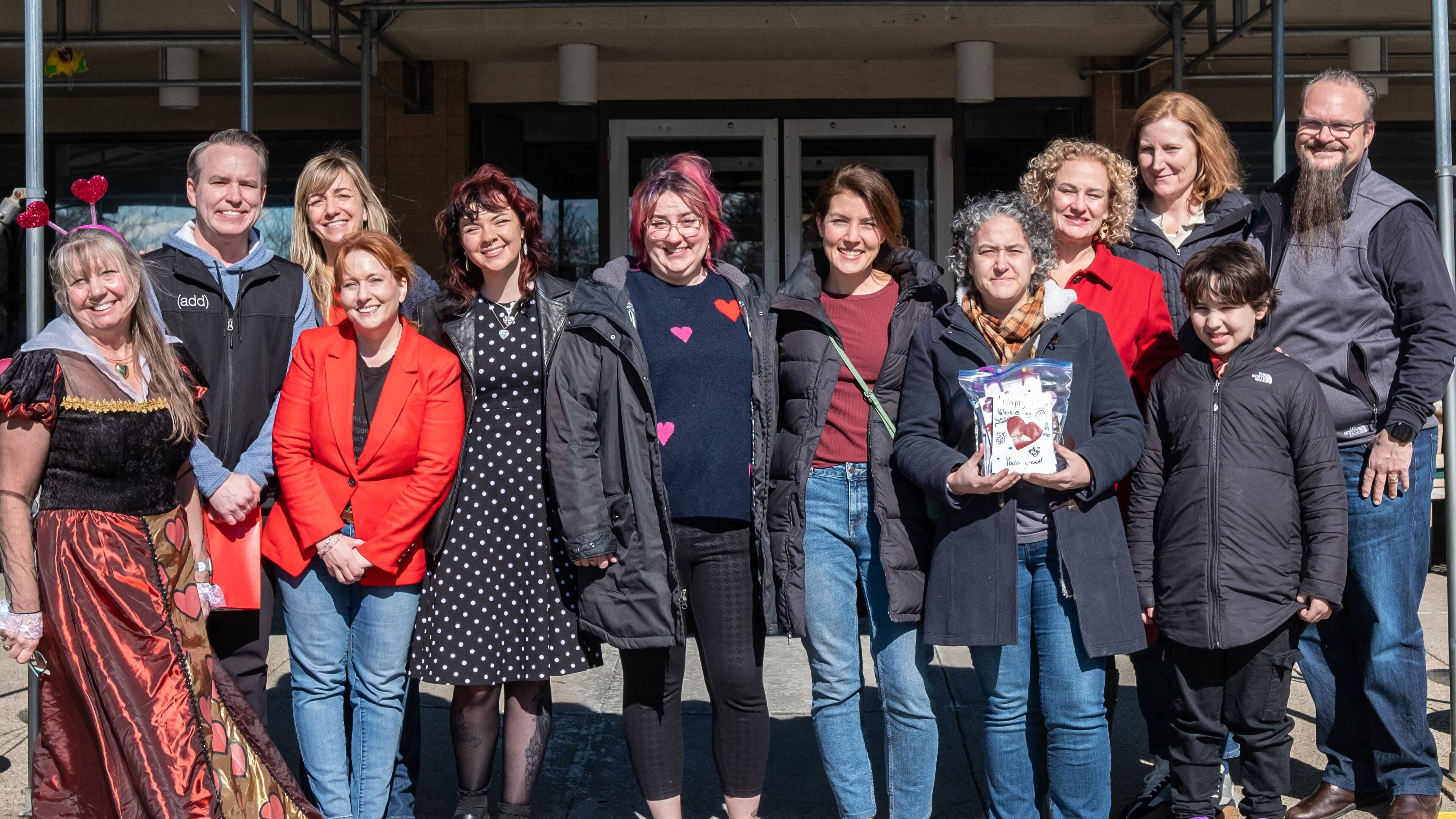  group of volunteers poses outside a building, dressed in festive and casual attire, celebrating a community Valentine’s Day event.