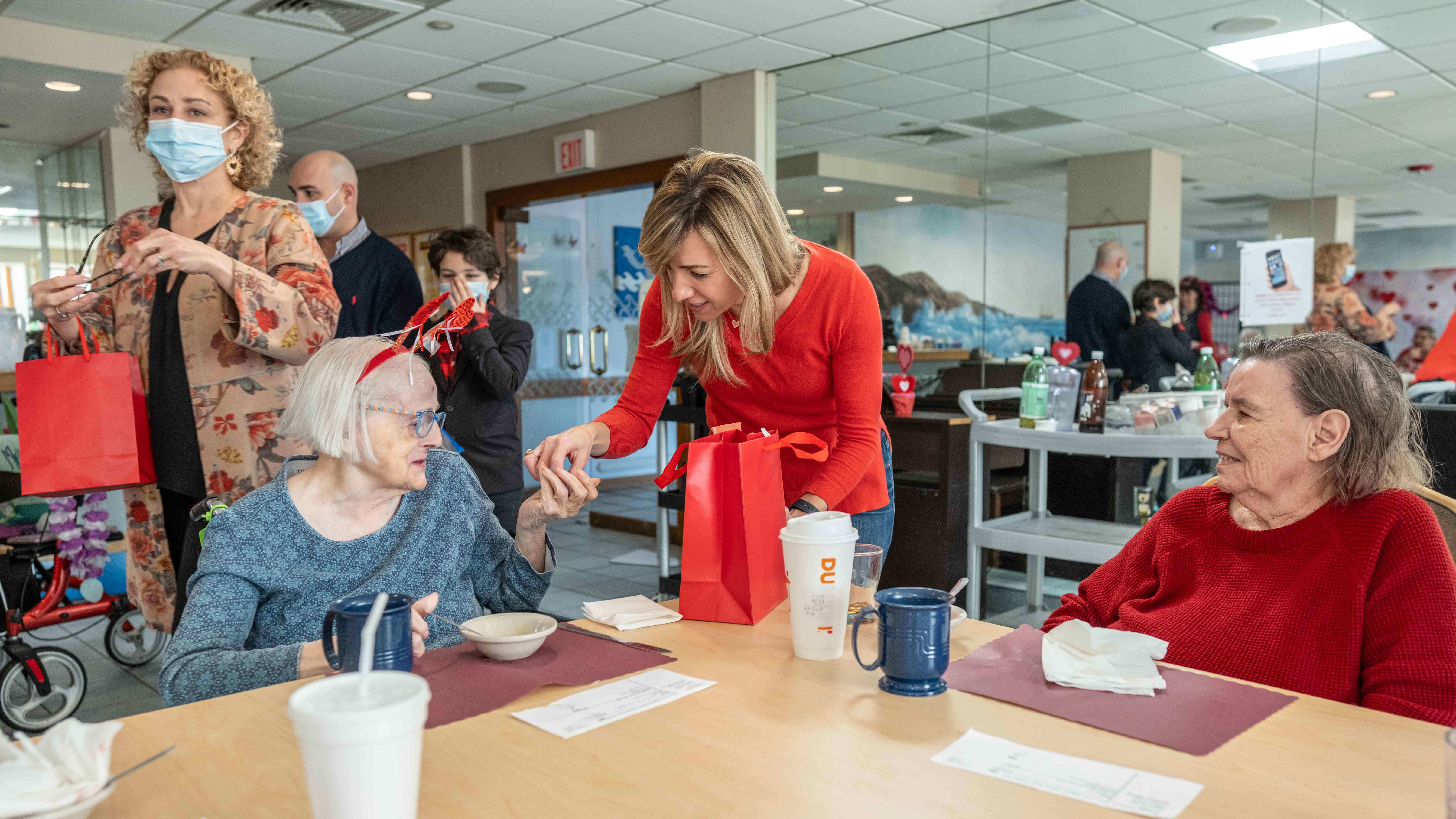 A volunteer in red hands a gift bag to an elderly woman wearing heart antennae at a Valentine’s Day event in a senior care facility.