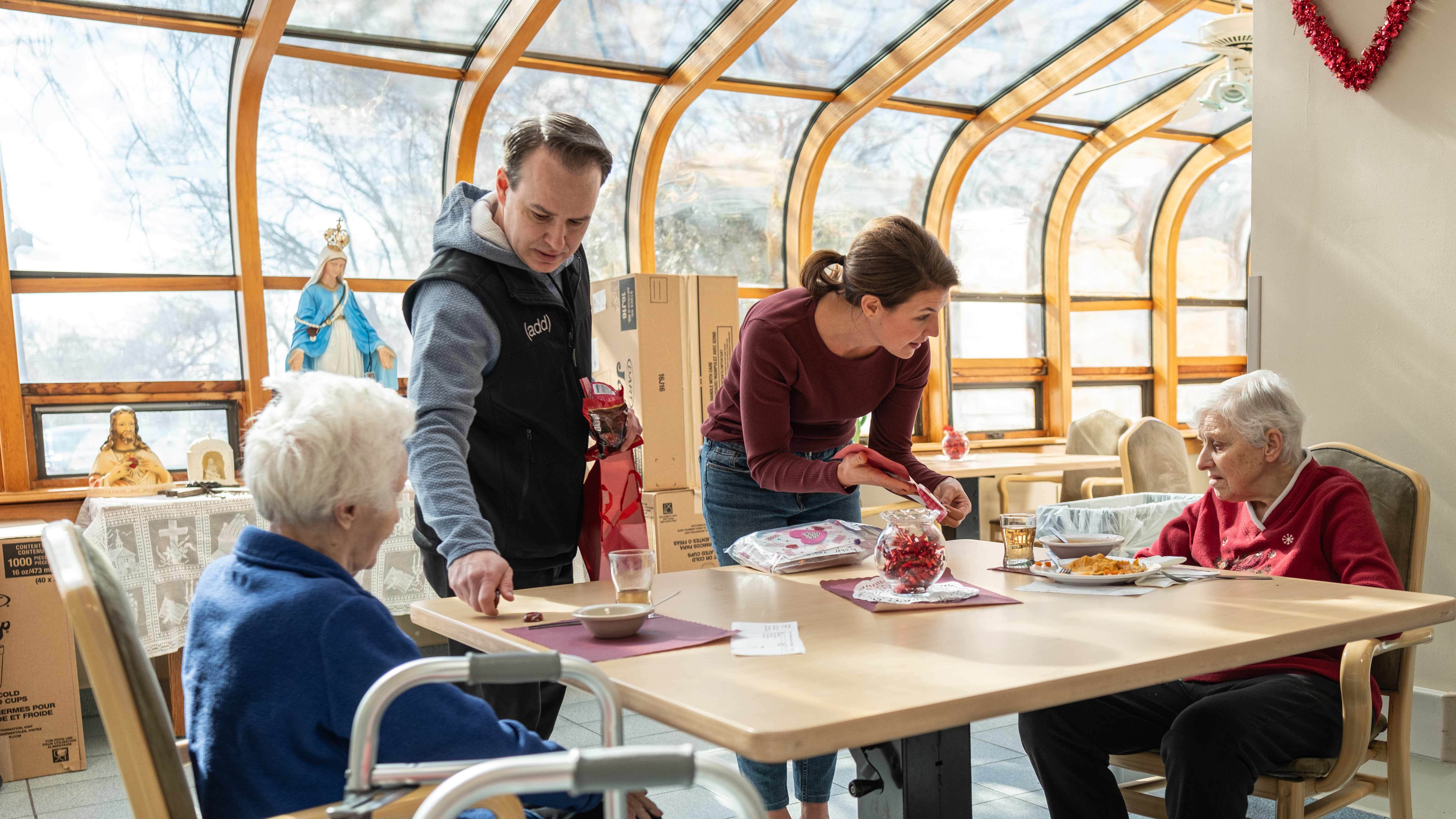 Two volunteers distribute Valentine’s Day treats to elderly residents in a sunlit room, decorated with festive red accents.