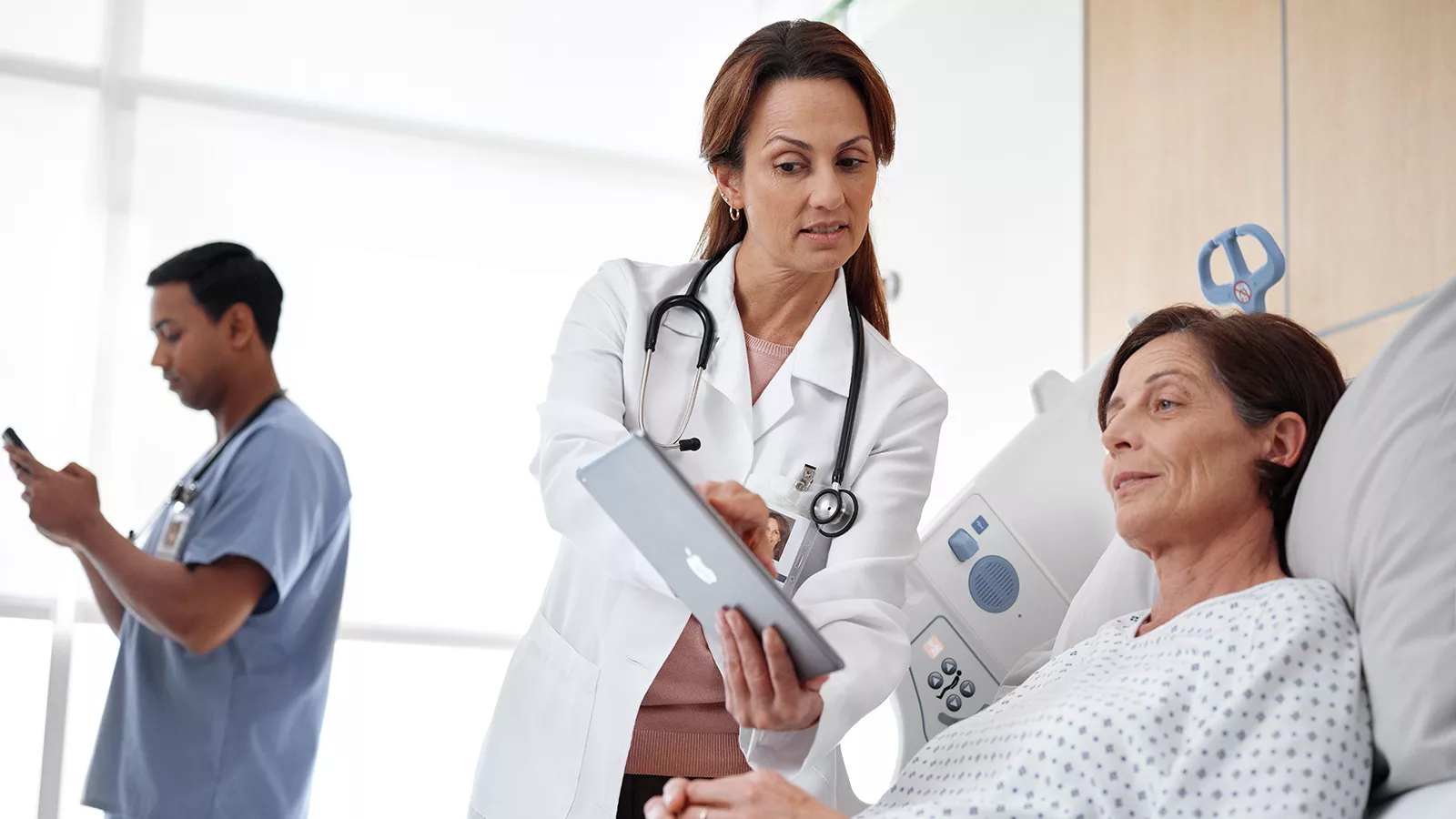 A female doctor in a white coat showing a tablet to a female patient in a hospital bed while a nurse uses a smartphone in the background.