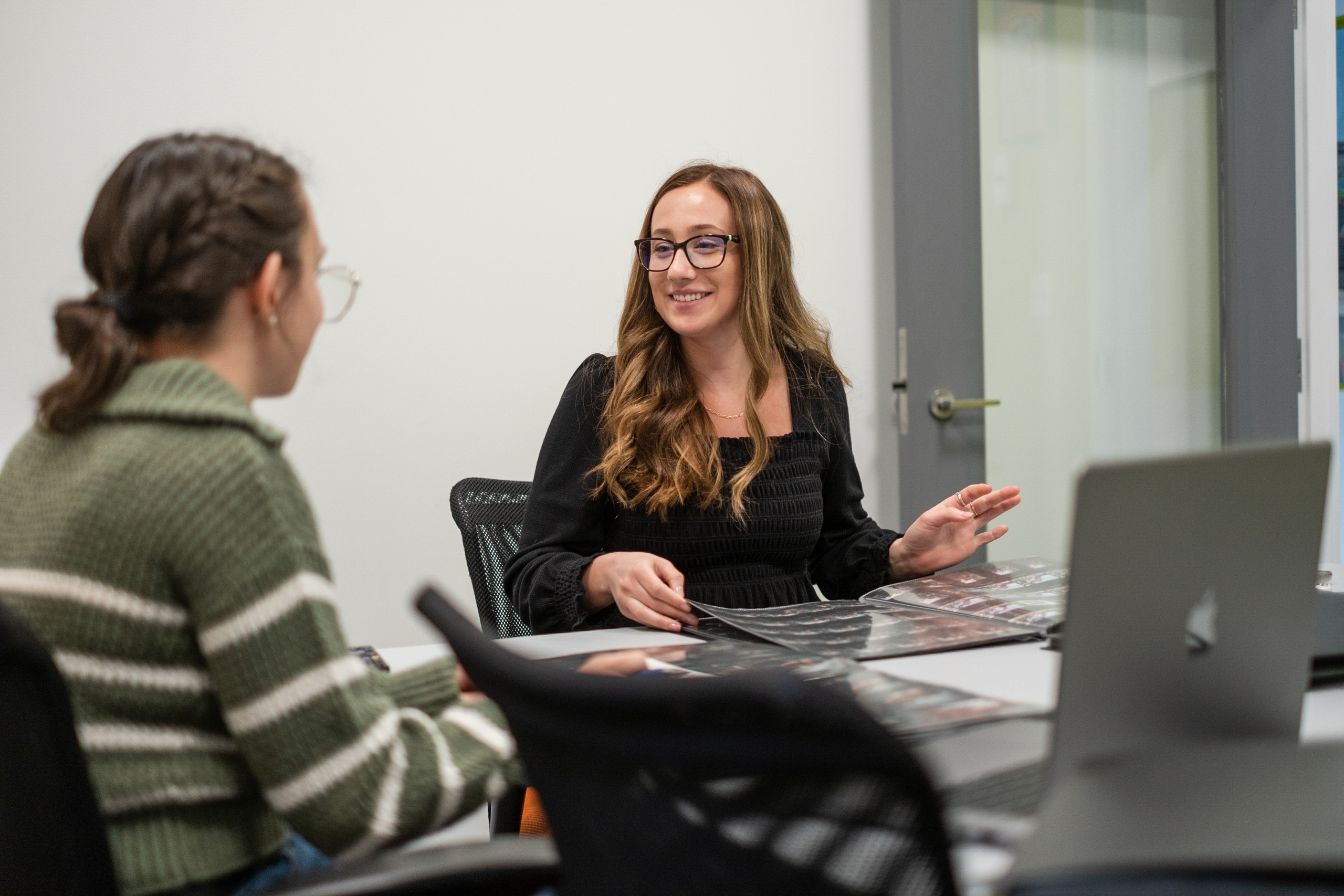  Two women sitting at a table in an office setting, discussing documents and a laptop, with one of them smiling.