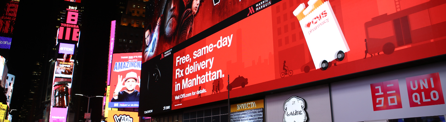 A large digital billboard in Times Square advertising CVS Pharmacy’s free same-day prescription delivery in Manhattan, with a red background and white text, alongside other brand advertisements.